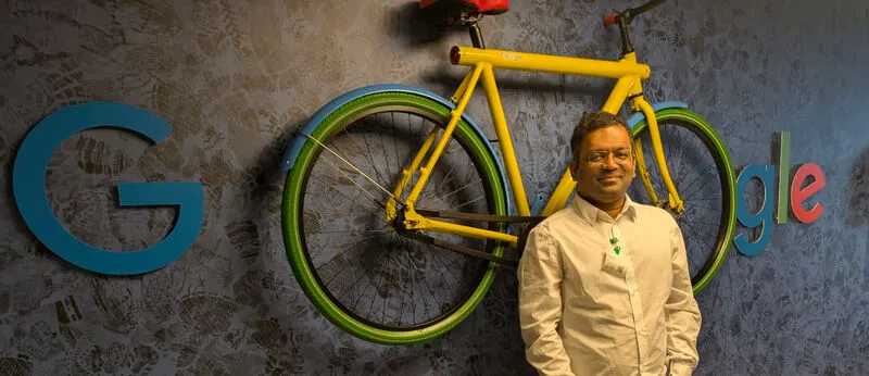 Ravindra Shinde standing in front of the Google logo wall, beside a yellow Google bike, at the Amsterdam office.